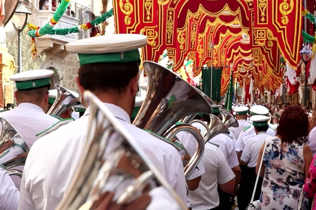 Reference photograph: A band marching in white caps beneath festa banners, beside the paper model rebuilt from it
